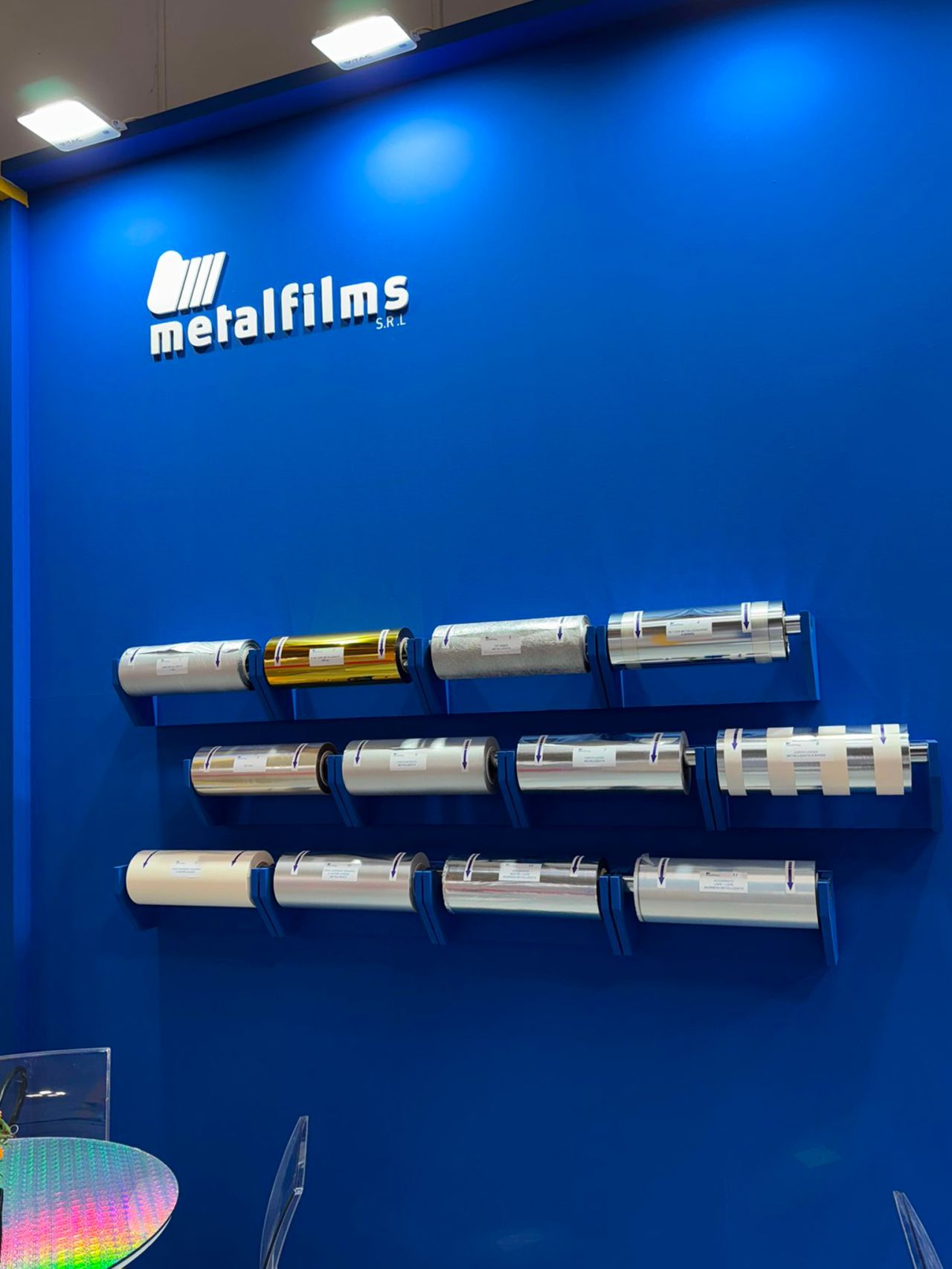 Parete espositiva Metalfilms con bobine di materiali tecnici metallizzati e riflettenti, presentati in fiera su fondo blu.