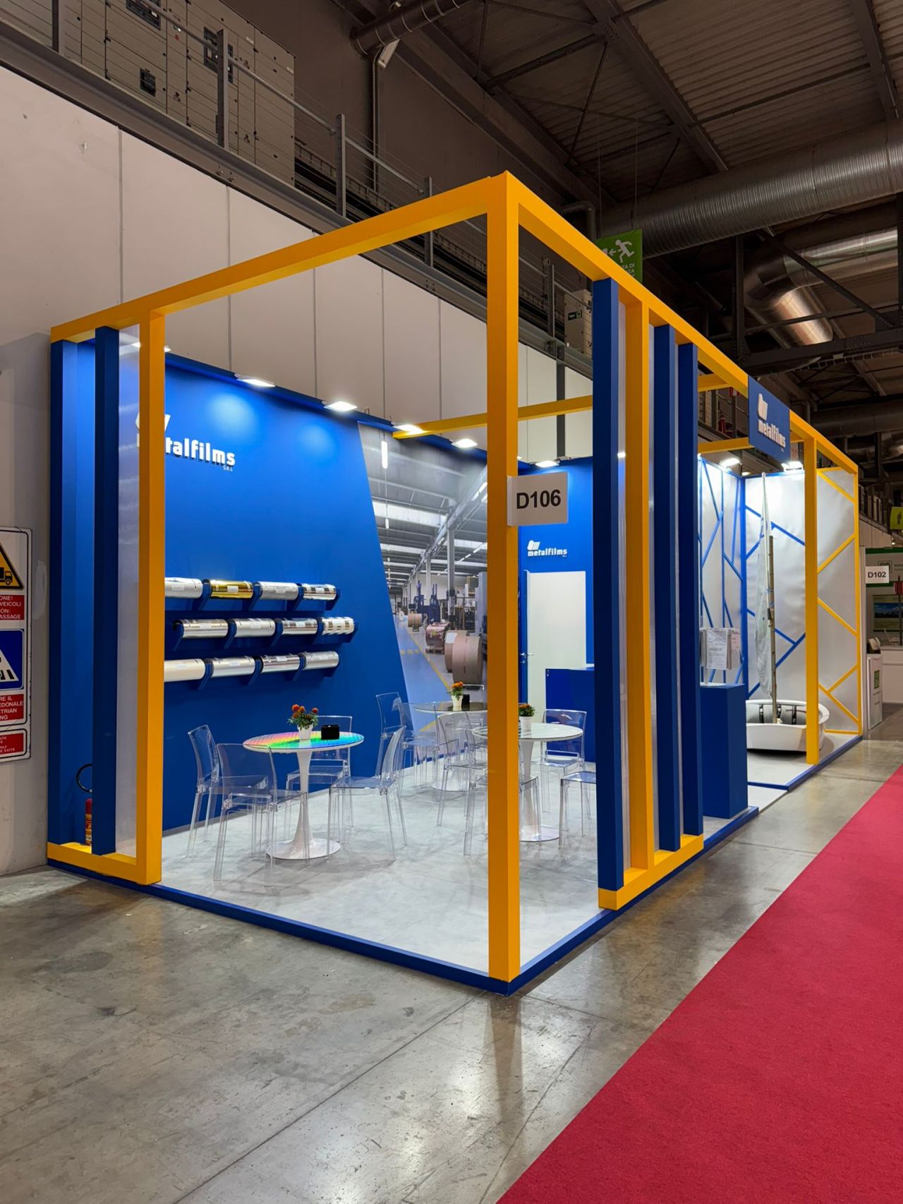Vista laterale dello stand Metalfilms allestito in fiera, con struttura gialla e blu, tavoli trasparenti e rotoli di materiale tecnico esposti.
