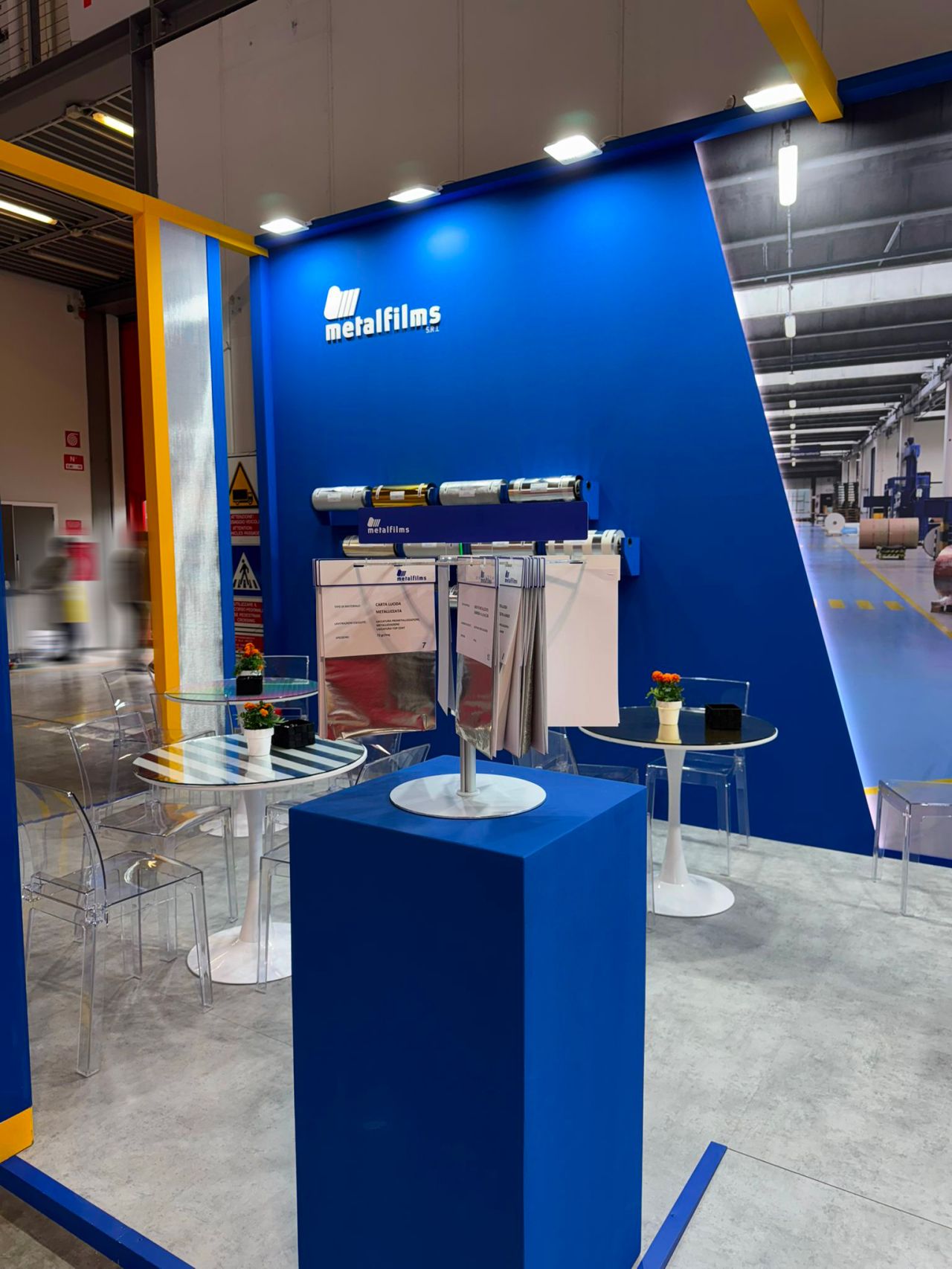 Area informativa dello stand Metalfilms con tavoli trasparenti, brochure e campioni di materiali tecnici esposti in fiera.