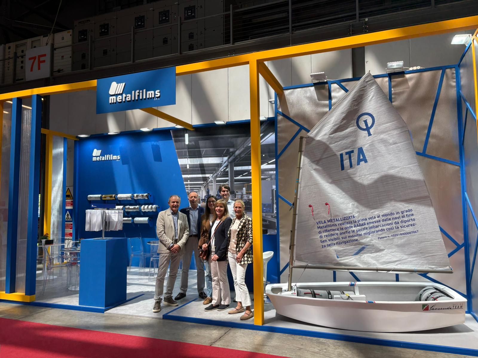 Il team Metalfilms in posa davanti allo stand fieristico con materiali tecnici e vela metallizzata esposta, durante evento di settore.